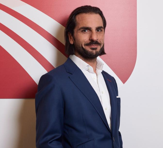 The image features a man wearing a blue suit standing against a red background. The man has a beard and is smiling at the camera. The text in the image suggests that there might be an update for the Bakkt cryptocurrency exchange, which could potentially affect users of the exchange who hold certain bank accounts or are interested in neobanking/fintech solutions.