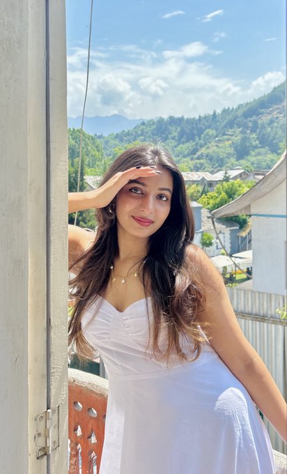 The image features a young woman wearing a white dress, posing for a photo while leaning against a wall or railing. She has her hand resting on her cheek, giving off an air of confidence and style. The background includes a mountain range under a blue sky, adding depth and context to the scene. The post caption reads "Finally doxxed after 6 years Hey there!!", which is likely meant as a playful response to the woman's image being shared or posted on social media platforms.

The woman in the white dress appears to be the main subject of the photo, with her outfit and pose drawing attention. Her hand resting on her cheek adds an element of casual elegance to the overall composition. The mountain range in the background provides a stunning backdrop for this image, enhancing its visual appeal and making it more memorable.