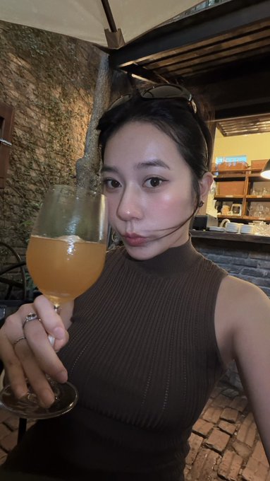 A woman in a brown ribbed sleeveless top takes a selfie while holding a glass of orange-colored beverage in what appears to be an outdoor evening setting.