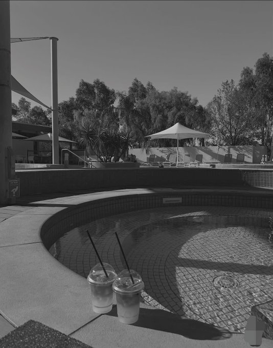 A black and white photo of two iced drinks sitting on the edge of an empty curved pool, with lounge chairs, a white umbrella, and palm trees visible in the background.