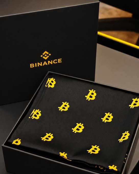A black gift box with the Binance logo opens to reveal a black fabric item (appears to be a face mask or bandana) printed with yellow Bitcoin symbols.