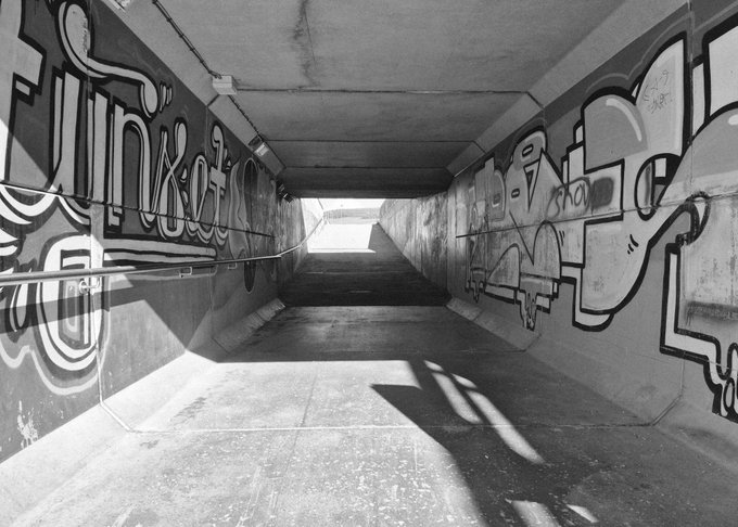 A black and white photograph shows the inside of a graffiti-covered concrete pedestrian tunnel or underpass.
