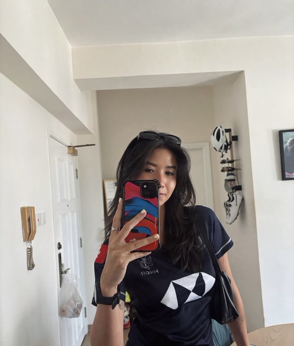 A mirror selfie of a young woman wearing sunglasses on her head and a dark blue shirt with a geometric logo, holding a phone with a red and blue case in a hallway.