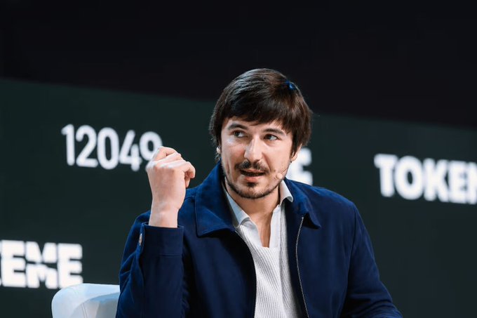 A man with dark hair and a beard, wearing a navy blue jacket over a white shirt, speaks animatedly at a conference with "TOKENIZE" displayed on a screen behind him, as tokenized real-world assets on Robinhood Chain surge to $70 million.