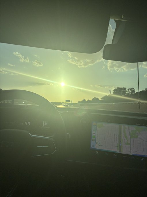A photograph taken from inside a car on a highway, showing the sun low in the sky, the dashboard displaying a speed of 59 mph, and a navigation map on the central screen.