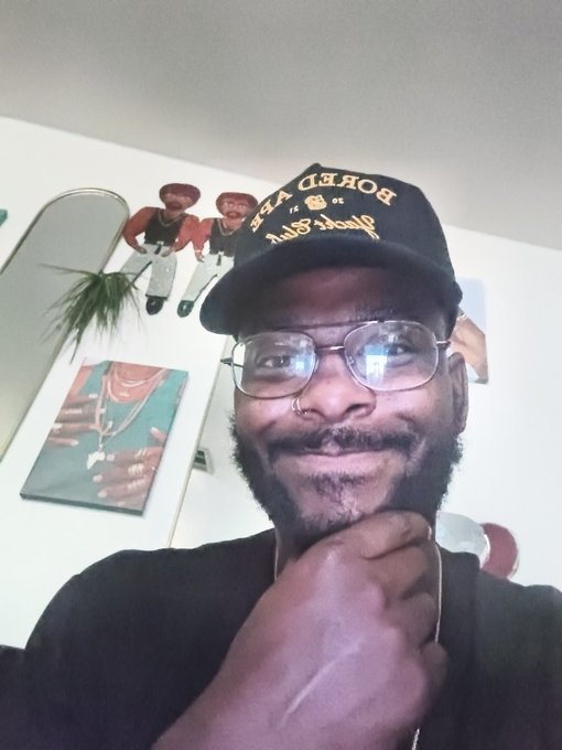 The image features a man wearing glasses, a black shirt, and a baseball cap with "Day 535" written on it. The man has his hand over his mouth as if he's about to make a funny face or joke. There is also an image of a person in a hat hanging above the man's head. The caption reads "#Day535 #apecoin A Supporting #yuga @apecoin Blessings ApE".