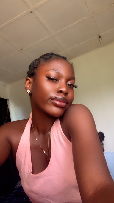 A young woman with braided hair, dramatic false eyelashes, and a pink halter top takes a selfie from a low angle.