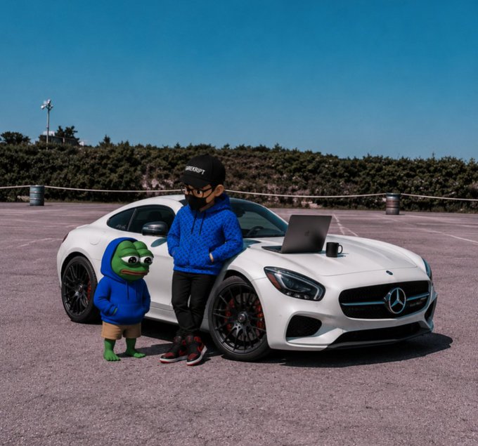 A photograph of a person wearing a black beanie, sunglasses, and a blue puffer jacket leaning against a white Mercedes-AMG car with a laptop and mug on the hood, accompanied by a Pepe the Frog character in a matching blue hoodie.
