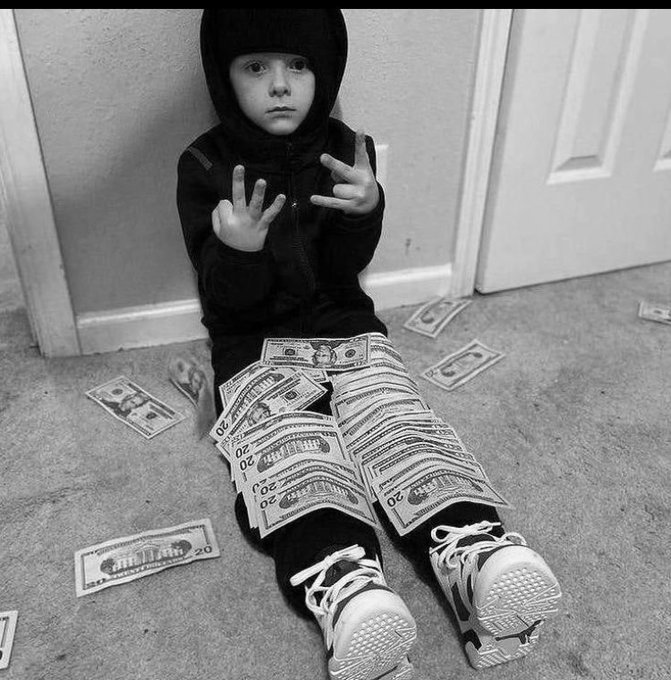 A black-and-white photo shows a young child in a hooded jacket sitting on the floor, covered with scattered twenty-dollar bills and making hand gestures.