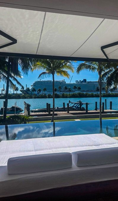 A view from beneath a canopy shows white lounge cushions in the foreground, a pool, and a large cruise ship docked across the water lined with palm trees.