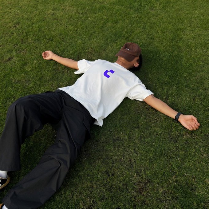 A person lies sprawled face-down on grass wearing a white t-shirt featuring the purple "C" logo of Crypto.com, perfectly capturing the exhausted "survived another week in crypto" vibe.