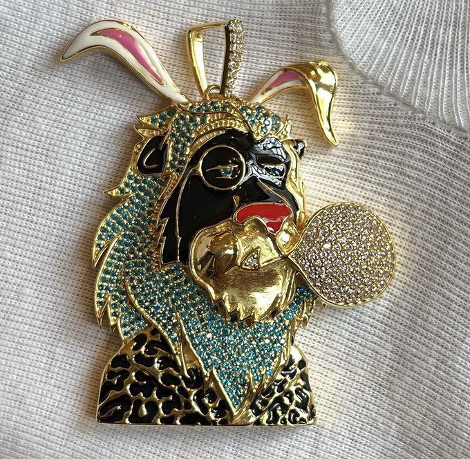 A gold-toned pendant featuring a goat-like figure with a hood, holding a diamond-encrusted heart, set against a white background.