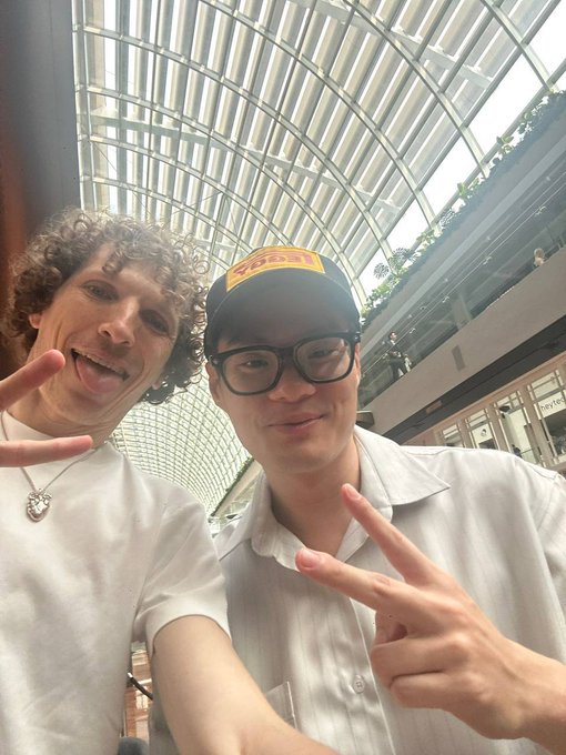 A selfie of two young men making peace signs in a shopping mall with a glass arched ceiling; one wears a white shirt with curly hair, and the other wears a red and yellow cap and black glasses.