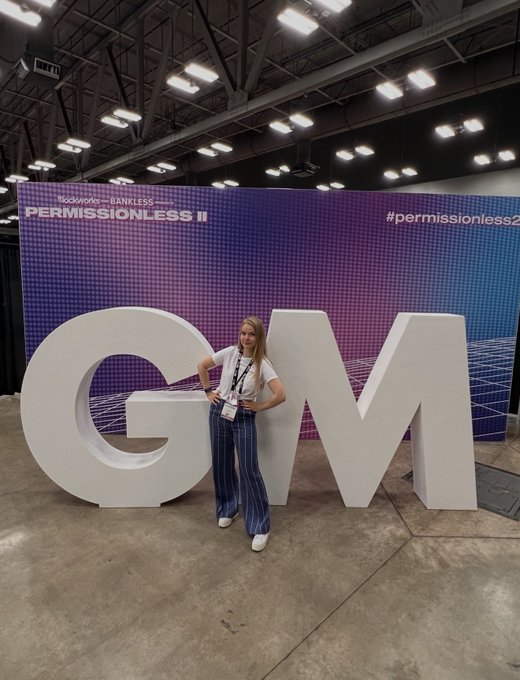 A woman poses in front of large white "GM" letters at a Permissionless II conference by Bankless, captioned to playfully reflect on the social media tradition of saying "GM" (good morning) as a community-building ritual.