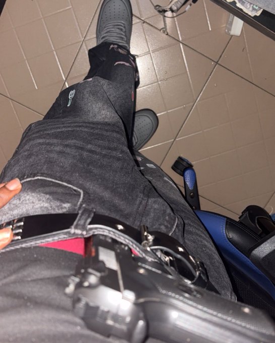 A first-person perspective photo looking down at a person wearing black jeans with decorative patterns and dark shoes, sitting on a blue chair with a black object in the foreground.