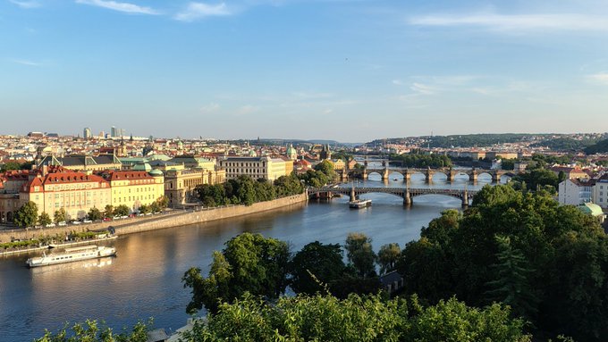 A sweeping summer view of Prague captures the sunlit Vltava River lined with historic buildings, with boats cruising beneath the city's iconic bridges, perfectly echoing the sentiment of Prague being the most beautiful city in summer.