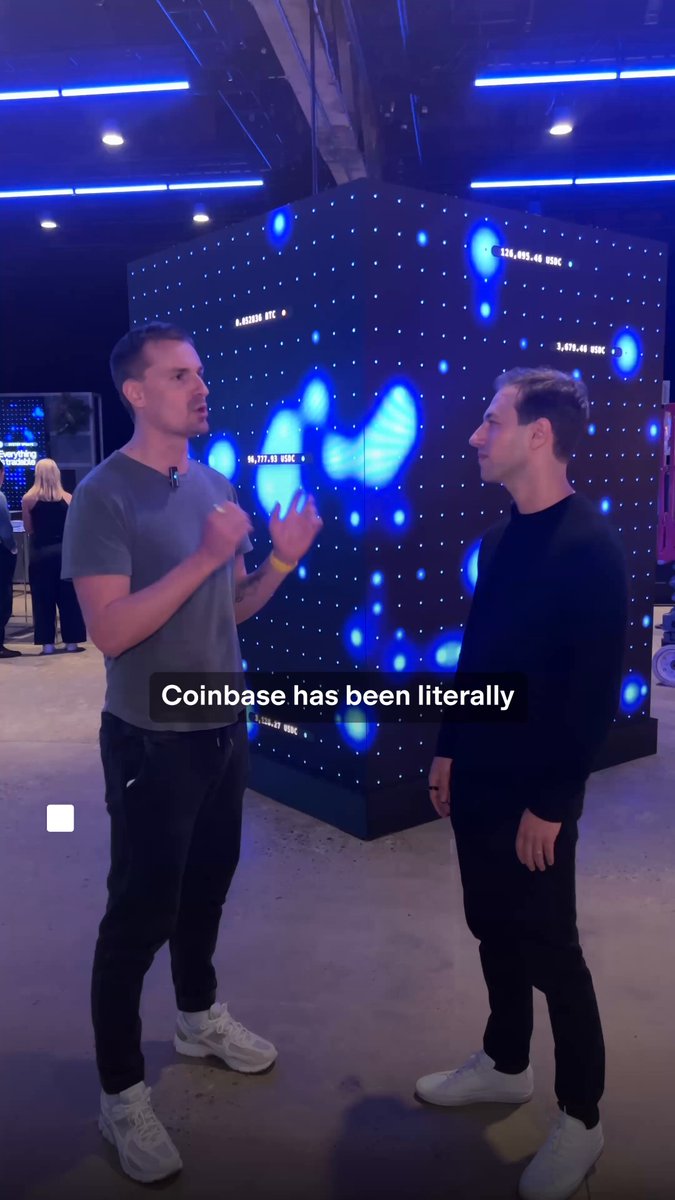 The image shows two men standing next to each other, engaged in a conversation. One man is wearing a gray shirt while the other has his back turned towards him. They are both looking at something off-camera, possibly discussing a recent video or photo they have seen on social media. The background features a large screen displaying blue dots and lines, which could be related to the coinbase platform or an advertisement for it.