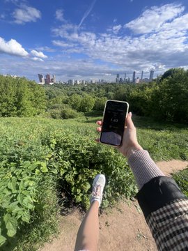 The image shows a person holding up their phone, which has an app open displaying the time as 12:00 pm (noon). This suggests that they are taking advantage of the first summer day with good weather conditions for outdoor activities or capturing photos. The presence of trees and greenery in the background further supports this idea, indicating that it is a sunny day outside.