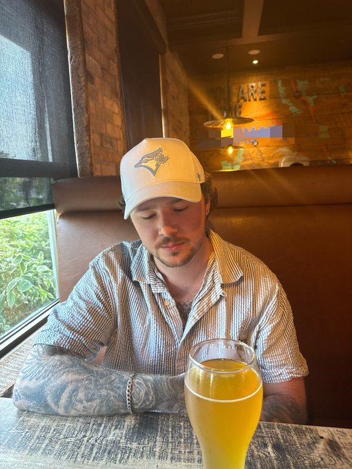 A man in a Toronto Blue Jays cap and striped shirt sits in a restaurant booth, looking down beside a glass of orange drink. The post says “GM Tuesday,” turning the casual morning greeting into a relaxed start-of-the-week moment.