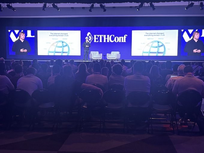 The image shows an event taking place at a conference center with a large screen displaying the name "ethcon" along with two speakers standing behind a podium, one of whom is wearing a suit. The audience consists of numerous people seated on chairs arranged in rows facing the stage where the speakers are presenting. The atmosphere appears to be professional and focused as attendees listen intently to the presentations.