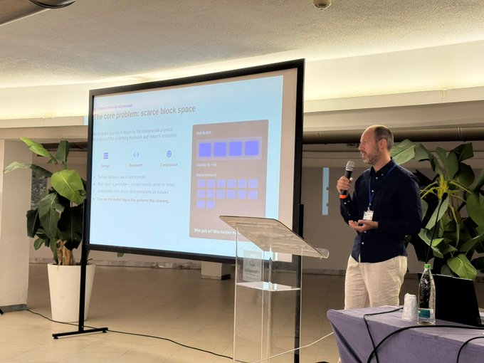 The image shows a man standing at a podium, giving a presentation with a large screen behind him displaying information about blockchain technology. The man appears to be wearing a blue shirt and white pants as he speaks into a microphone. There are potted plants in front of the stage, adding some greenery to the scene. A bottle is also visible on the table near the podium.