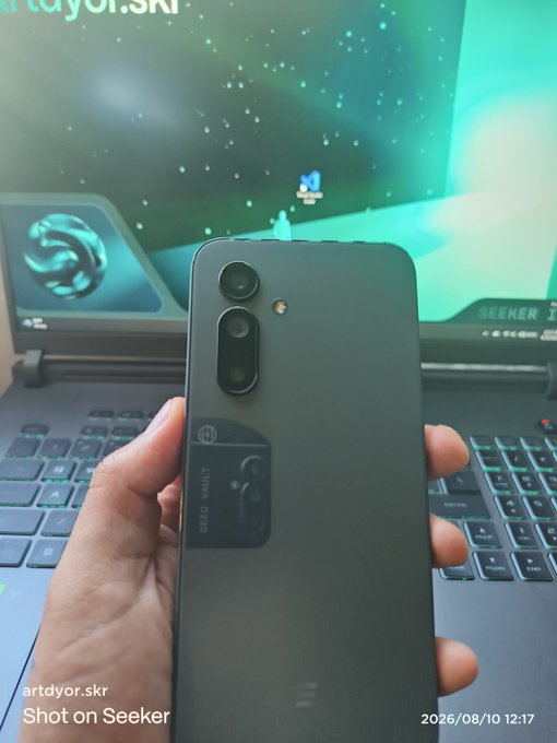 A person's hand holding a black smartphone in front of a laptop screen, with a watermark reading 'artdyor.skr Shot on Seeker 2026/08/10 12:17' at the bottom.