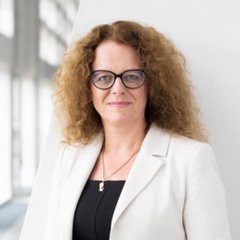 A professional headshot of a woman with curly blonde hair wearing glasses and a white blazer over a black top.