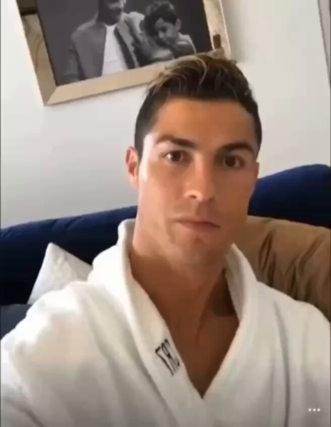 The image features a man wearing a white robe or bathrobe while sitting on a couch. He appears to be posing for a photo with a serious expression as he looks directly at the camera. The background of the image includes a picture frame, suggesting that this might be a home setting where the photo was taken.