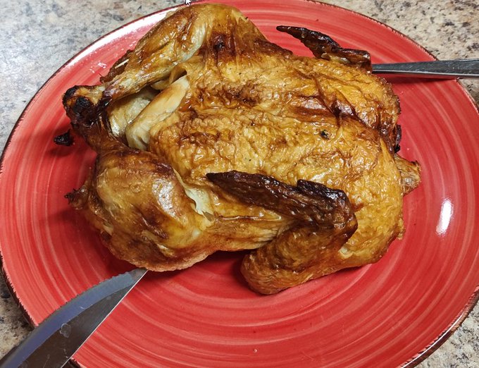 The image features a roasted chicken sitting on top of a red plate, which is placed on a table. There are two knives resting next to the plate - one closer to the left side and another towards the right side. The background appears to be a kitchen counter or dining table with some other items visible but not directly related to the main focus of the image.