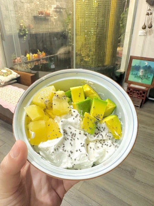 A hand holds a bowl containing yogurt topped with yellow and green fruit pieces (likely mango and avocado) sprinkled with chia seeds, with a cat visible in the background near a window.