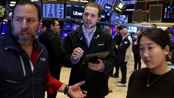Traders work on the floor of the New York Stock Exchange, with monitors in the background displaying 'Uber' and 'Prudential' tickers; a trader with 'BENNY' on his jacket speaks with others.