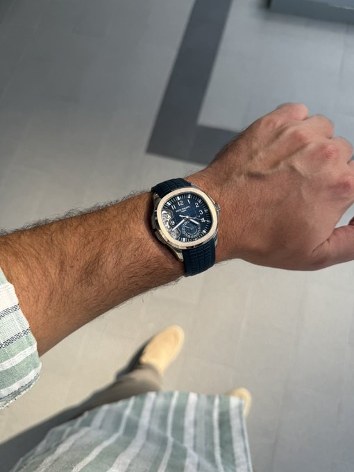 A first-person view of a man's wrist wearing an Audemars Piguet Royal Oak watch with a blue dial and black rubber strap, paired with beige shoes and striped pants.