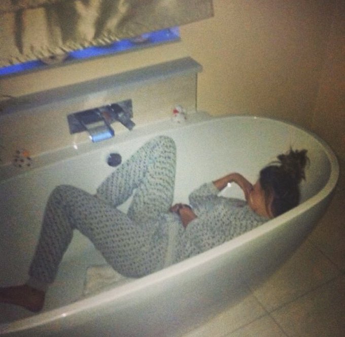 A woman with her hair tied up lies in a modern white bathtub, wearing patterned pajamas and looking at a smartphone.