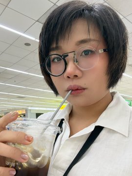 A person with short dark hair and round black-rimmed glasses drinks from a plastic cup with a straw, wearing a white collared shirt in what appears to be a brightly lit indoor setting.