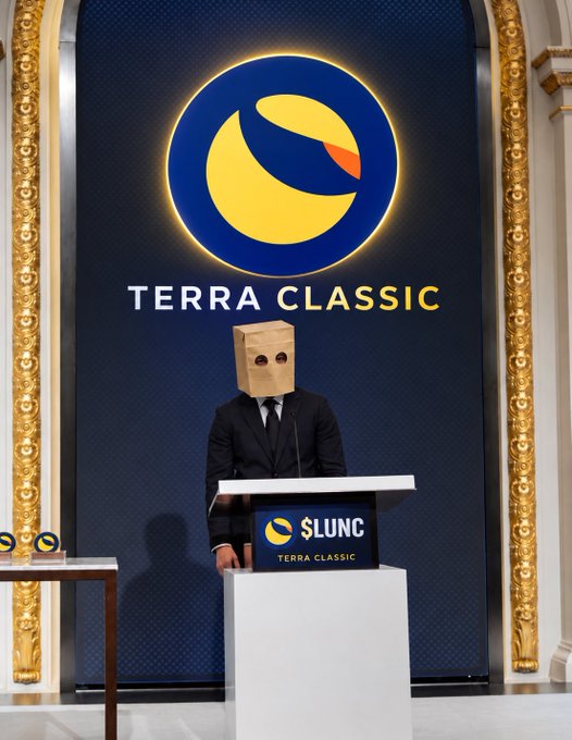 A figure in a suit and tie stands at a podium wearing a paper bag over their head, with the 'TERRA CLASSIC' logo displayed prominently on the backdrop and podium ($LUNC).