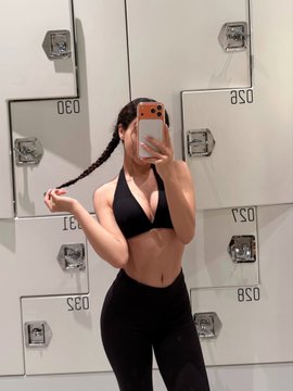 A mirror selfie of a woman in a black sports bra and leggings taking a photo with an orange smartphone in front of metal lockers numbered 030, 031, 057, and 058.