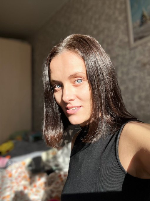 A selfie of a young woman with long brown hair and blue eyes, wearing a black tank top, taken in a sunlit room with a bed visible in the background.