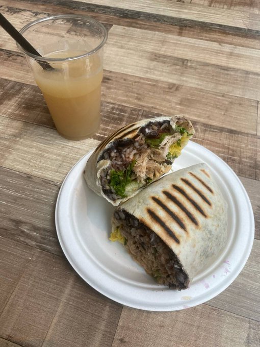 A plate holds a cut burrito with meat, beans, and lettuce next to a plastic cup of light orange juice on a wooden table.