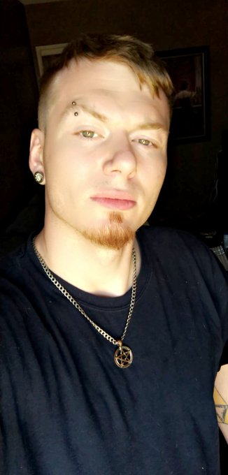 A close-up selfie of a young man with short blonde hair, a goatee, and facial piercings, wearing a black t-shirt and a silver chain necklace with a pendant.
