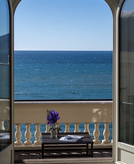 A view through an arched doorway onto a balcony with a white balustrade, overlooking a deep blue ocean under a clear sky, with a small table holding a vase of purple flowers.