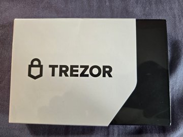 A white-and-black Trezor hardware wallet box rests on a dark surface, with the Trezor lock logo and the word “TREZOR” printed prominently.