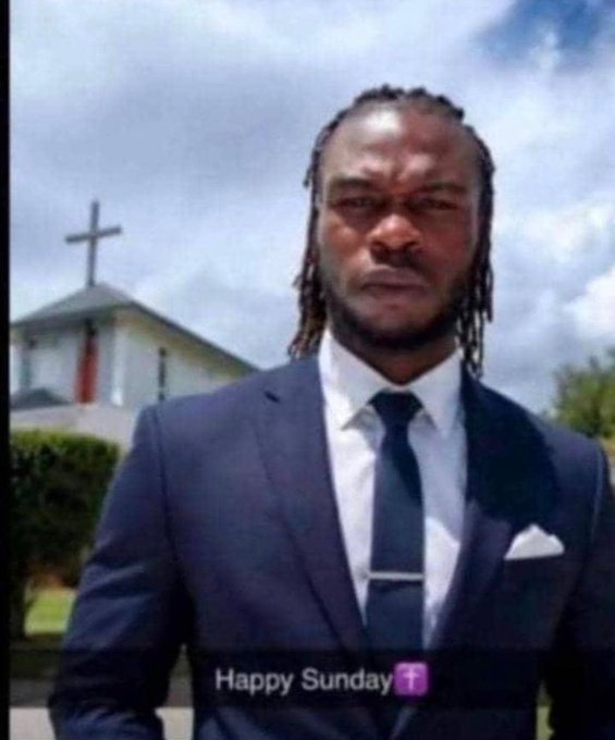 A man in a dark blue suit, white shirt, and black tie with braided hair stands outside a white church with a cross on the roof; the text 'Happy Sunday' with a cross emoji appears at the bottom.