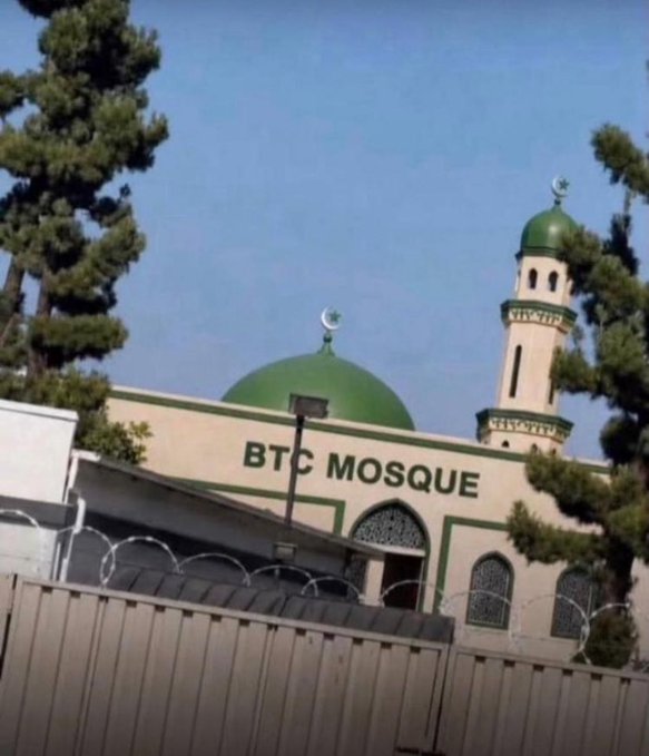 A mosque with a green dome and minaret is shown behind a fence with razor wire, with a sign on the building reading 'BTC MOSQUE'.