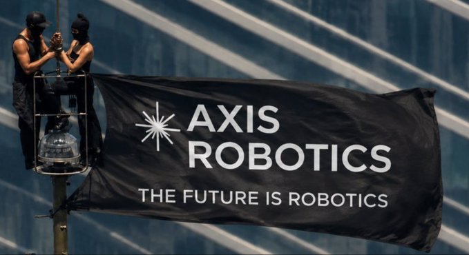 A promotional image showing two individuals installing a large black "AXIS ROBOTICS — THE FUTURE IS ROBOTICS" banner against a glass building, visually branding the robotics platform featured in the accompanying guide to Axis's tasks, yapping, and roles for new contributors.