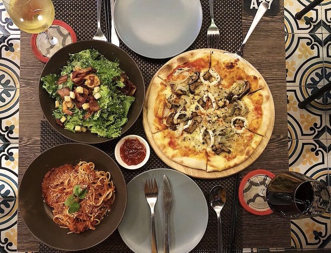A table spread featuring a salad with bacon, a pasta dish with marinara sauce, a whole pizza topped with mushrooms and onions, a glass of white wine, and a glass of red wine, viewed from above on a tiled surface.