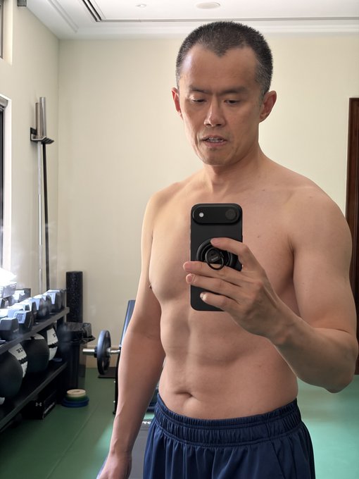 A shirtless, muscular middle-aged Asian man taking a mirror selfie with a smartphone in what appears to be a gym, with dumbbells and exercise equipment visible in the background.