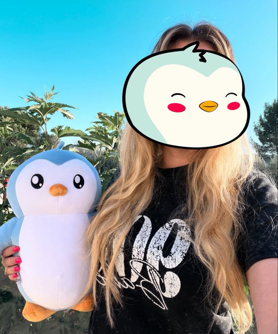 A woman with long blonde hair holds a plush penguin toy outdoors, her face obscured by a cartoon bird filter, while wearing a black shirt with white lettering.