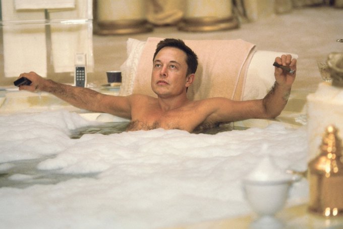 A man relaxes in a bubble bath with his arms outstretched, holding a phone in one hand and a cigar in the other, with a vintage telephone visible on the side.