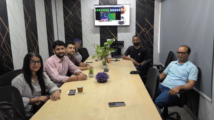 A group of five people sitting around a wooden conference table in a modern meeting room, with a monitor displaying a colorful interface mounted on the wall behind them.