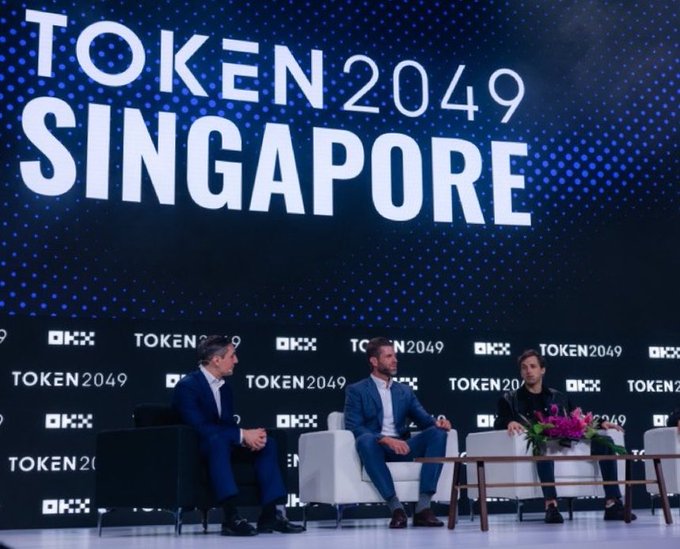 Three individuals sit on a stage at the TOKEN2049 Singapore conference, with the event's logo and "OKX" sponsor branding displayed on the large backdrop behind them. The image captures a panel discussion at the crypto conference, tying into the post's message of thanking Singapore while teasing the upcoming New York 2027 event.